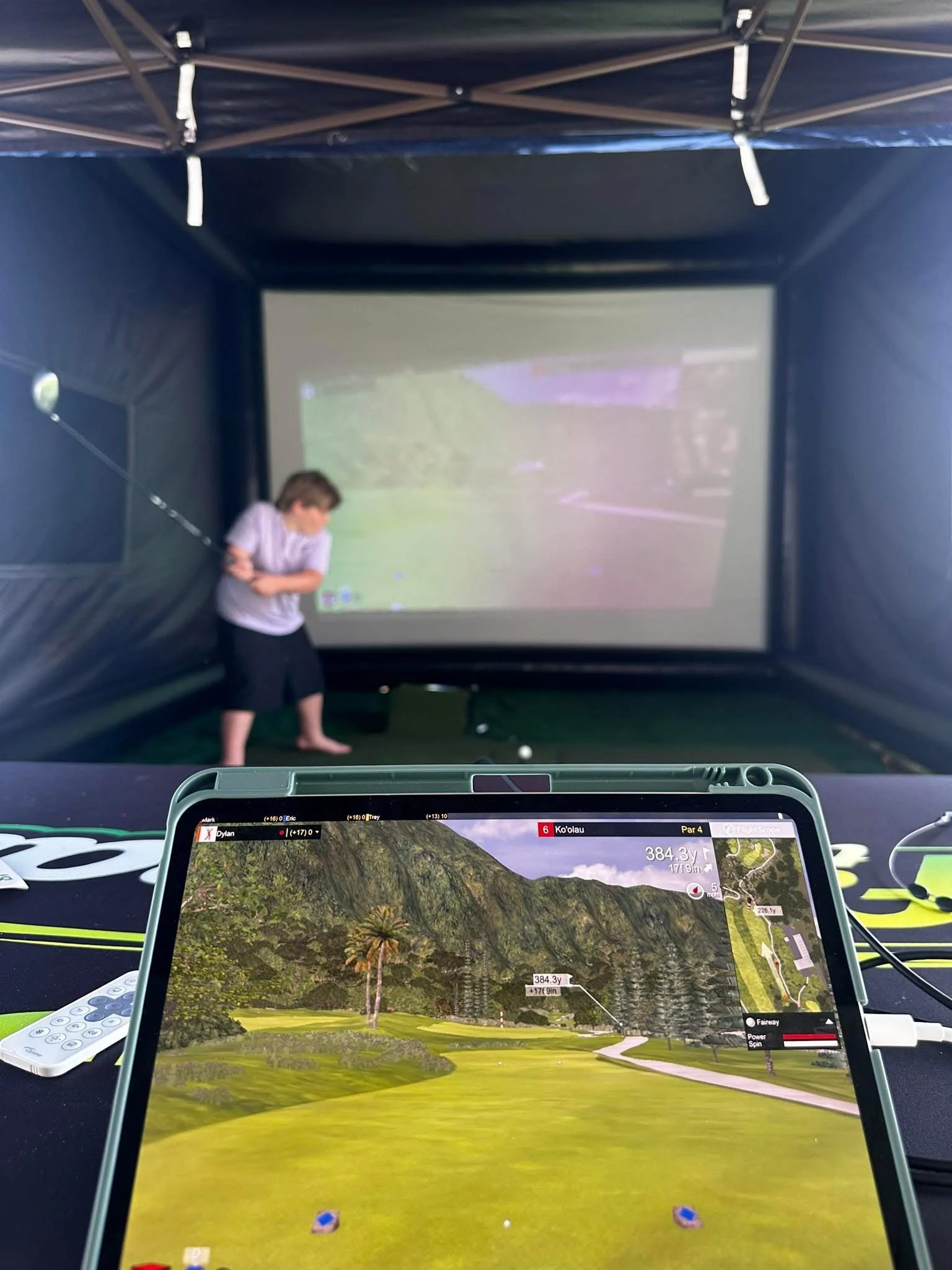 A golf simulation setup showing a screen with a course view, a person preparing to take a shot, and a tablet displaying a virtual golf course