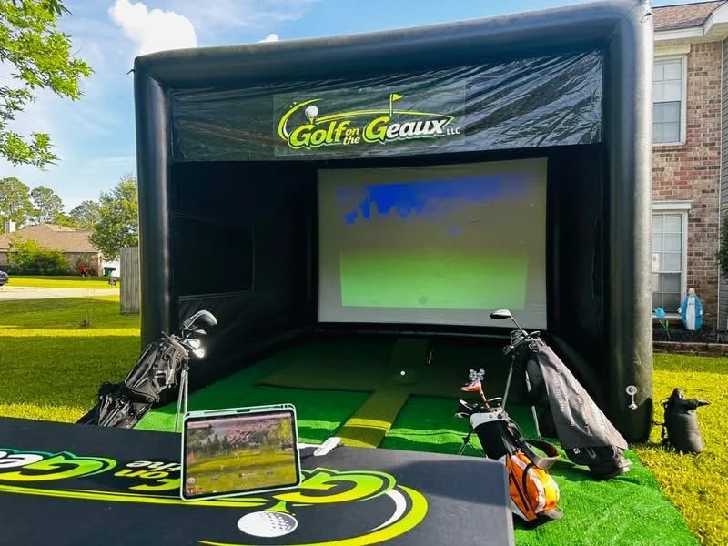 Outdoor golf simulator setup with a large screen, golf clubs, and golf bags, branded with 'Golf for the GEEAUX LLC' logo, on a grassy lawn in front of a house.