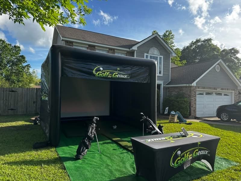 Inflatable golf simulator tent set up on a lawn in front of a house with a garage, with golf clubs and a table with golf branding.