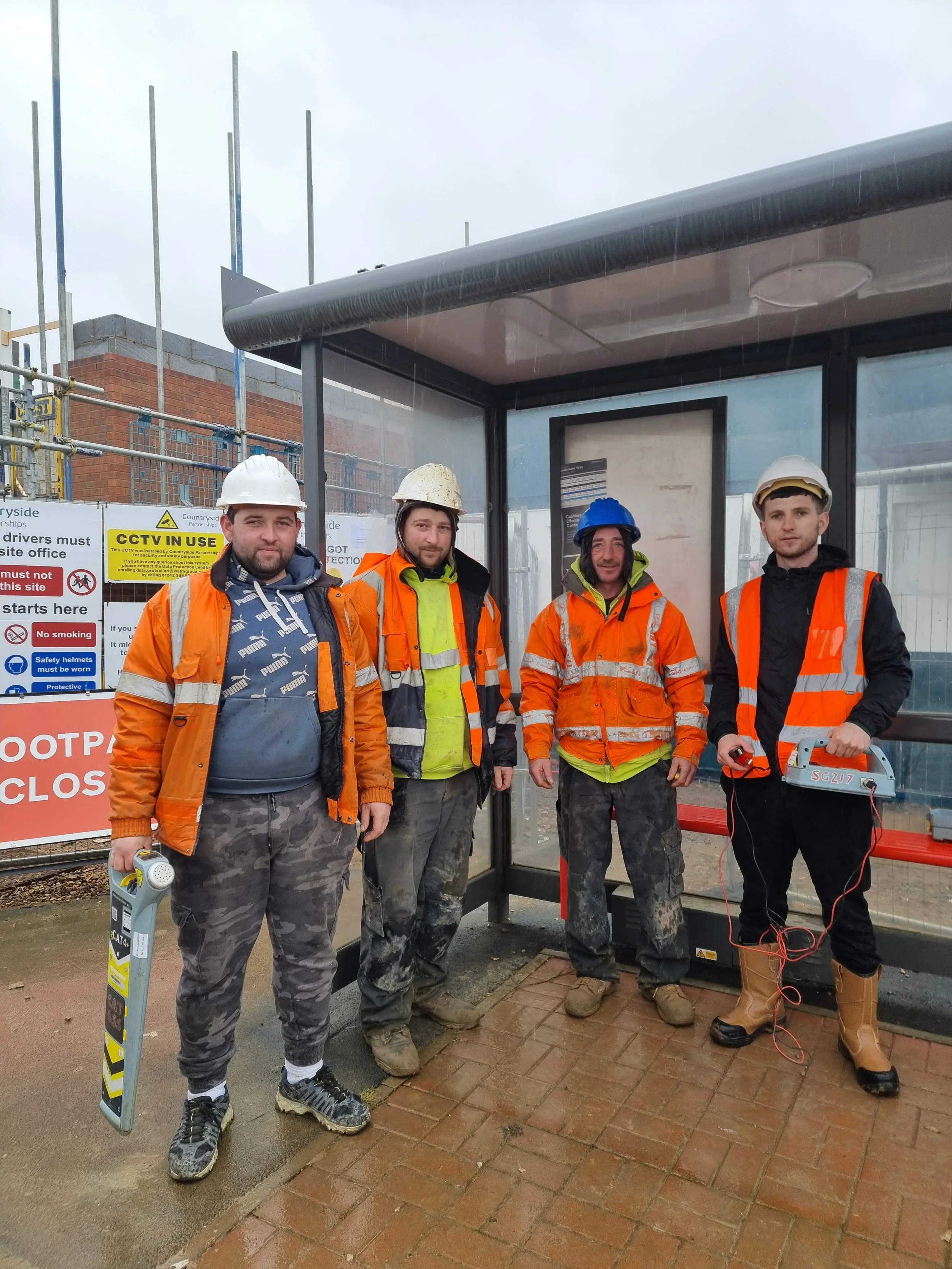 Four construction workers standing at a bus stop shelter on a rainy day. They are wearing helmets and high-visibility jackets. The person on the left is holding a device, and the person is holding a CAT and Genny.