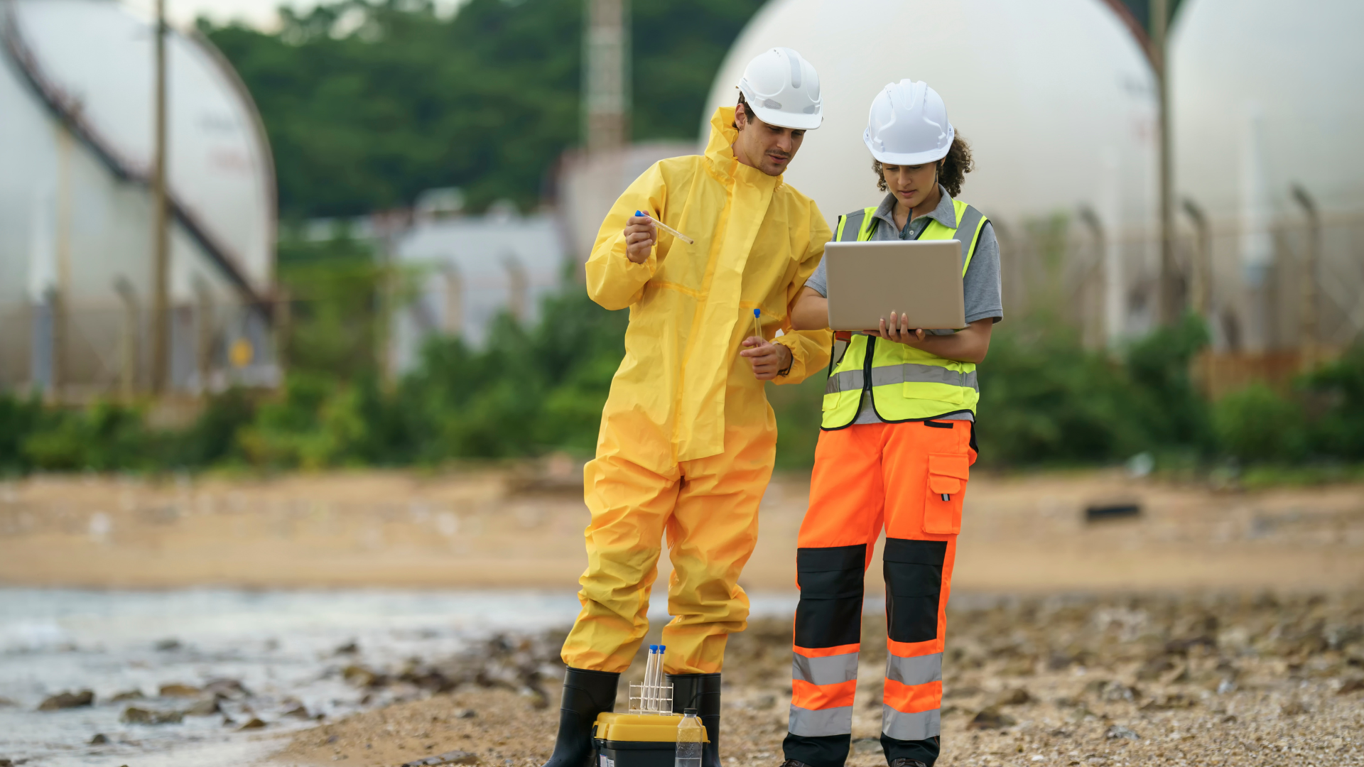 Two workers wearing safety helmets and bright safety clothing examining groundwater samples on a construction site with greenhouses in the background.