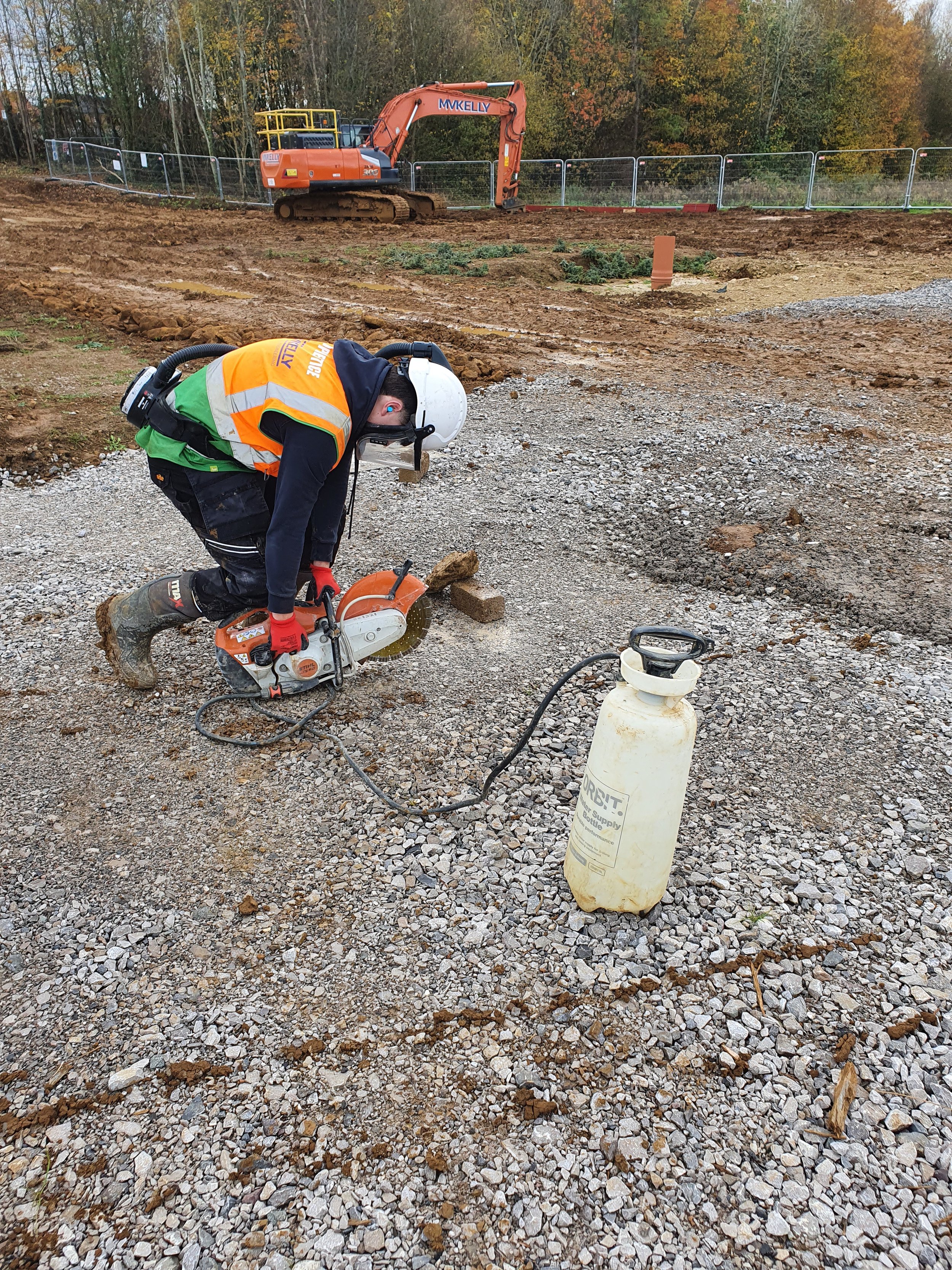 A construction worker wearing a white helmet, orange safety vest, green shirt, and red gloves is operating a small orange and white cut-off saw on a gravel surface at a construction site. In the background, there is an orange excavator with the label 'MVKELLY' and trees with fall foliage.