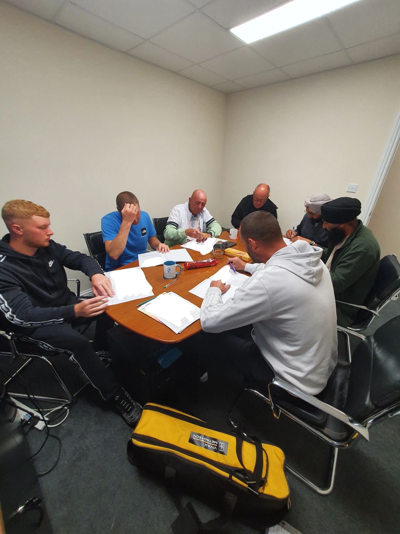 Six people sitting around a conference table in a meeting room, engaged in discussion and reviewing documents, with various mugs and supplies on the table.