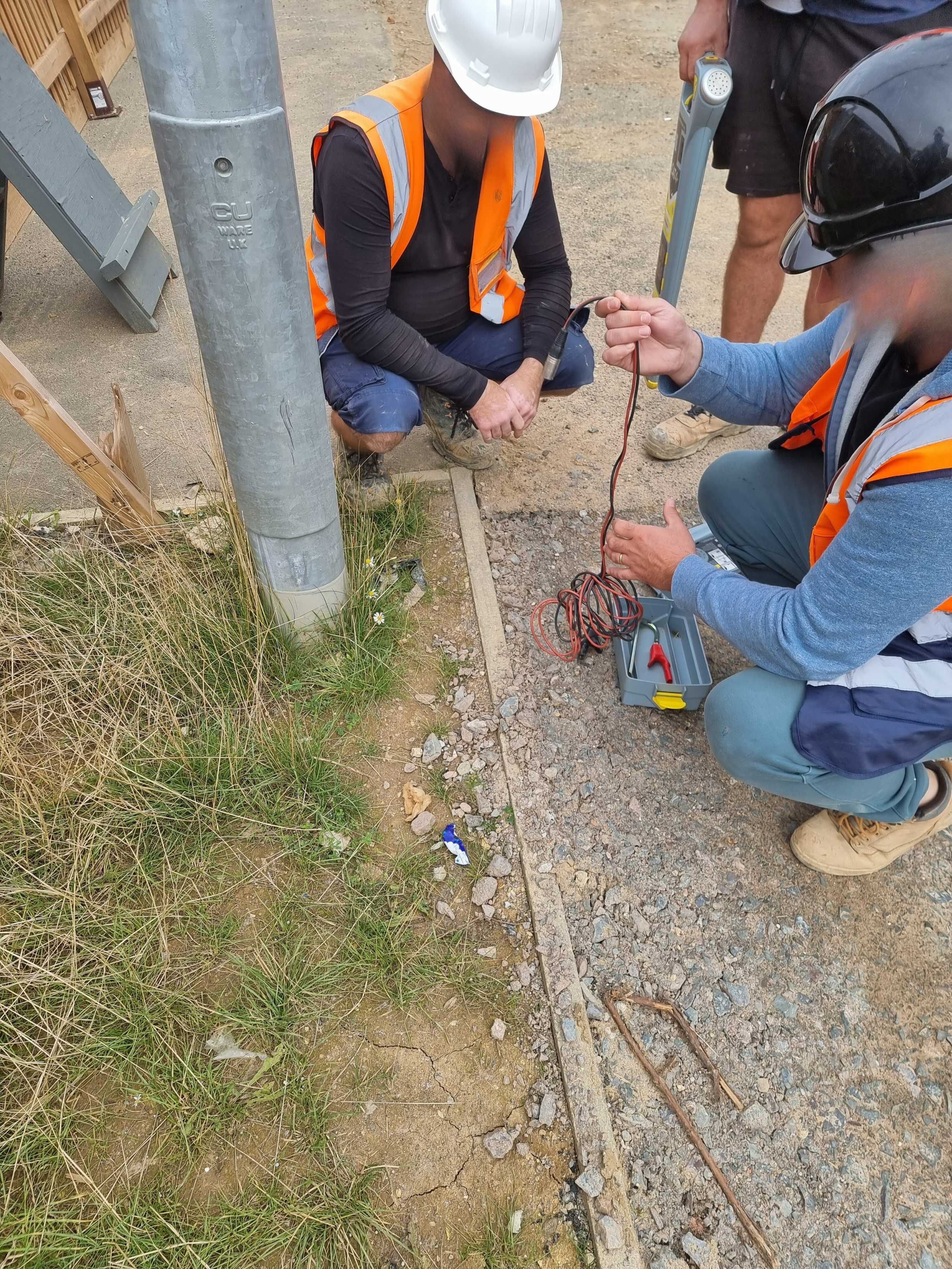 Two construction workers with safety helmets and vests working outside near a utility pole, using testing equipment with wires.