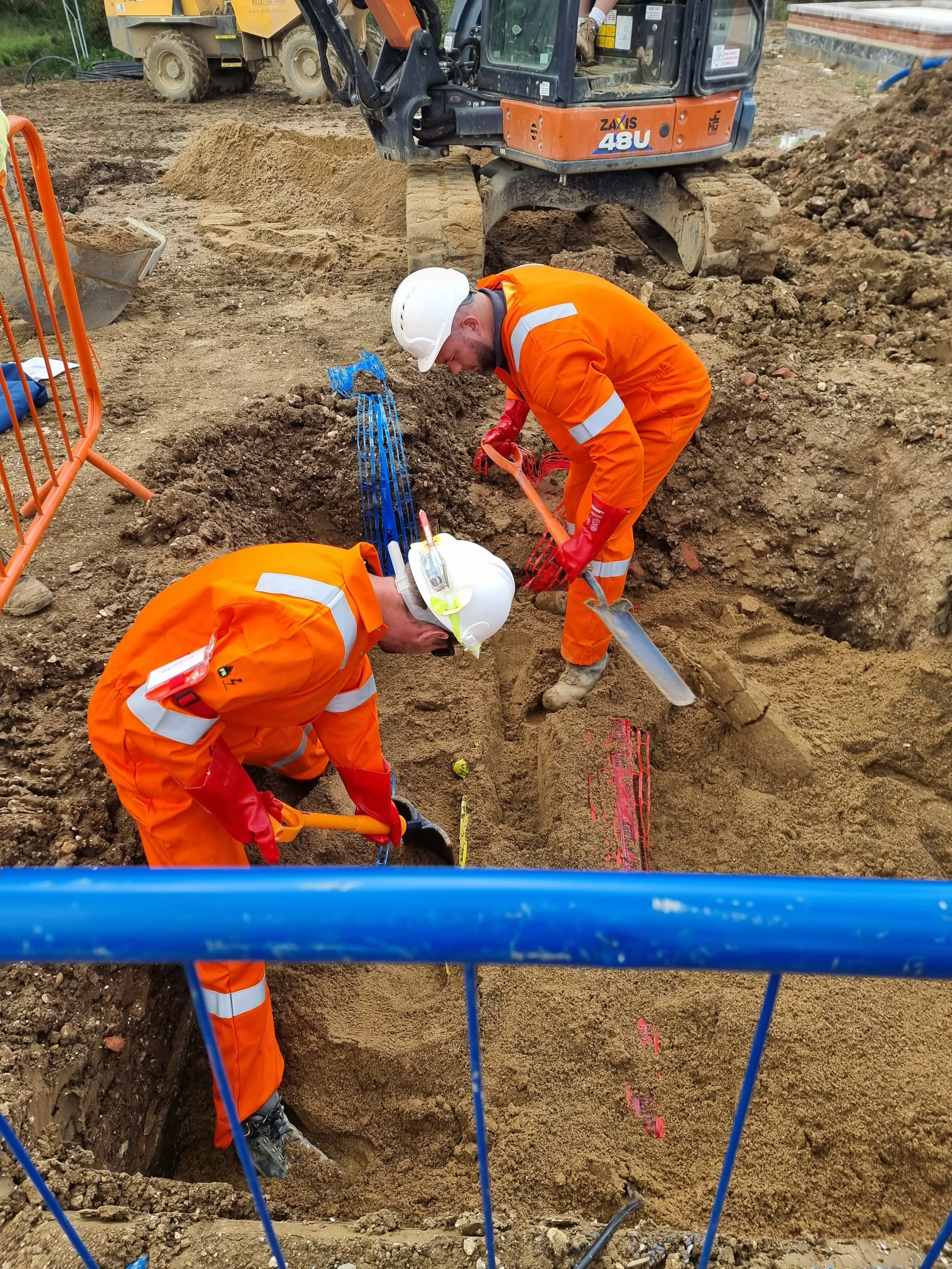 Construction workers in orange safety overalls and helmets working in a trench at a construction site. One worker is using a shovel, while another appears to be working on underground cables or pipes. Heavy machinery and construction materials are visible in the background.