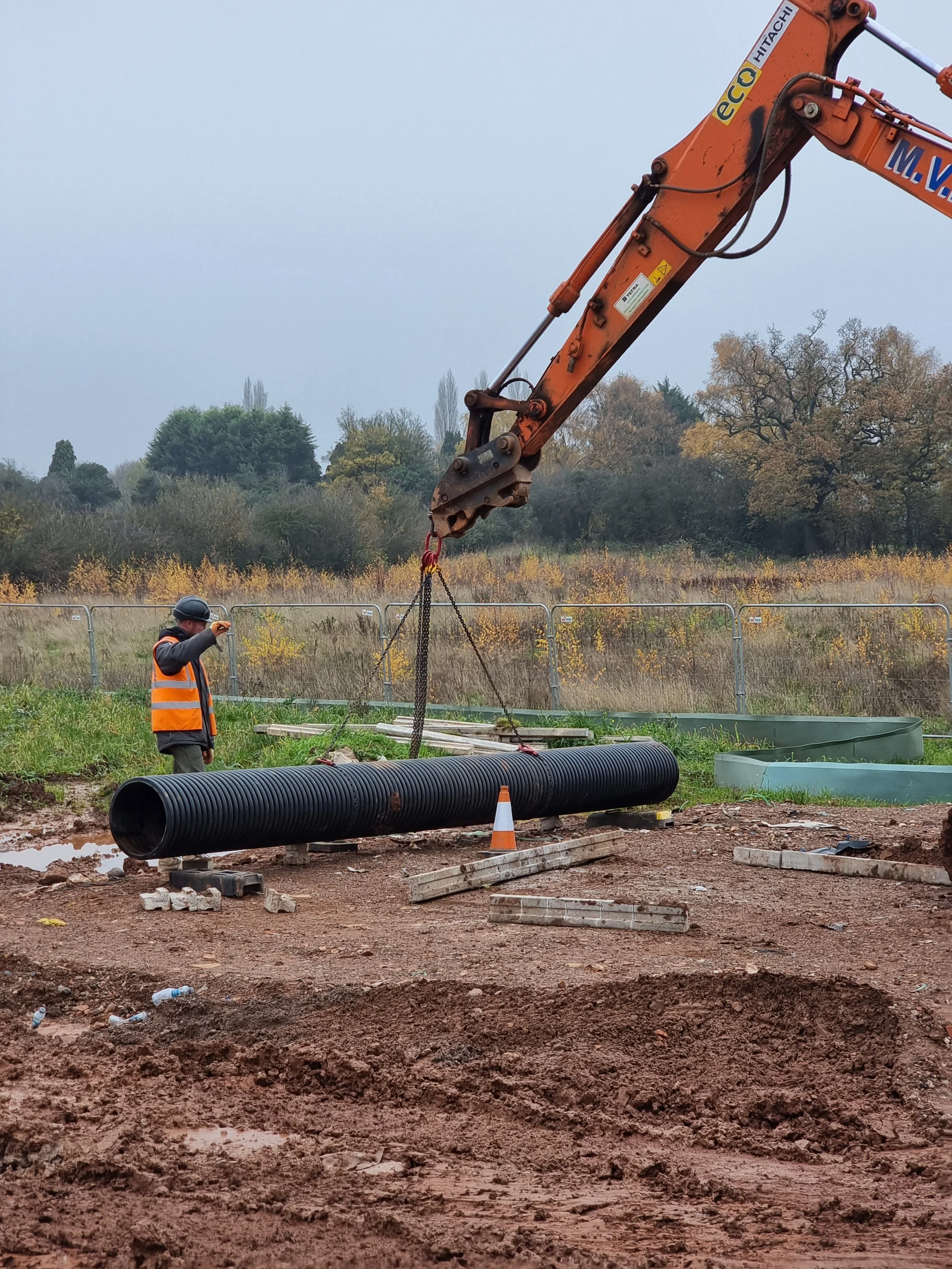 Construction site with a large black pipe on the ground, a worker in a safety vest and helmet, an excavator lifting a chain, and a fence in the background with trees and cloudy sky.