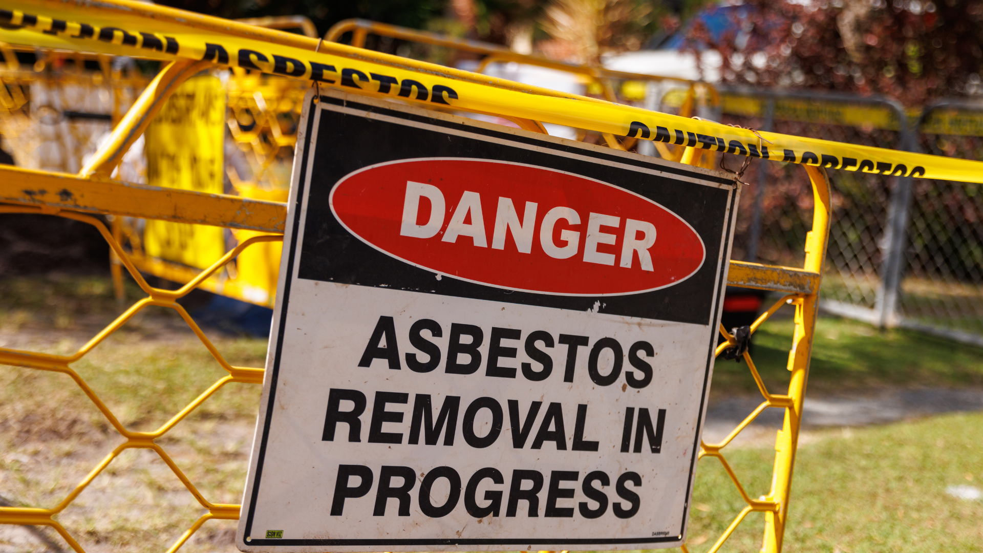 Yellow construction barrier with a warning sign that reads 'Danger Asbestos Removal in Progress' in front of a chain-link fence and trees.
