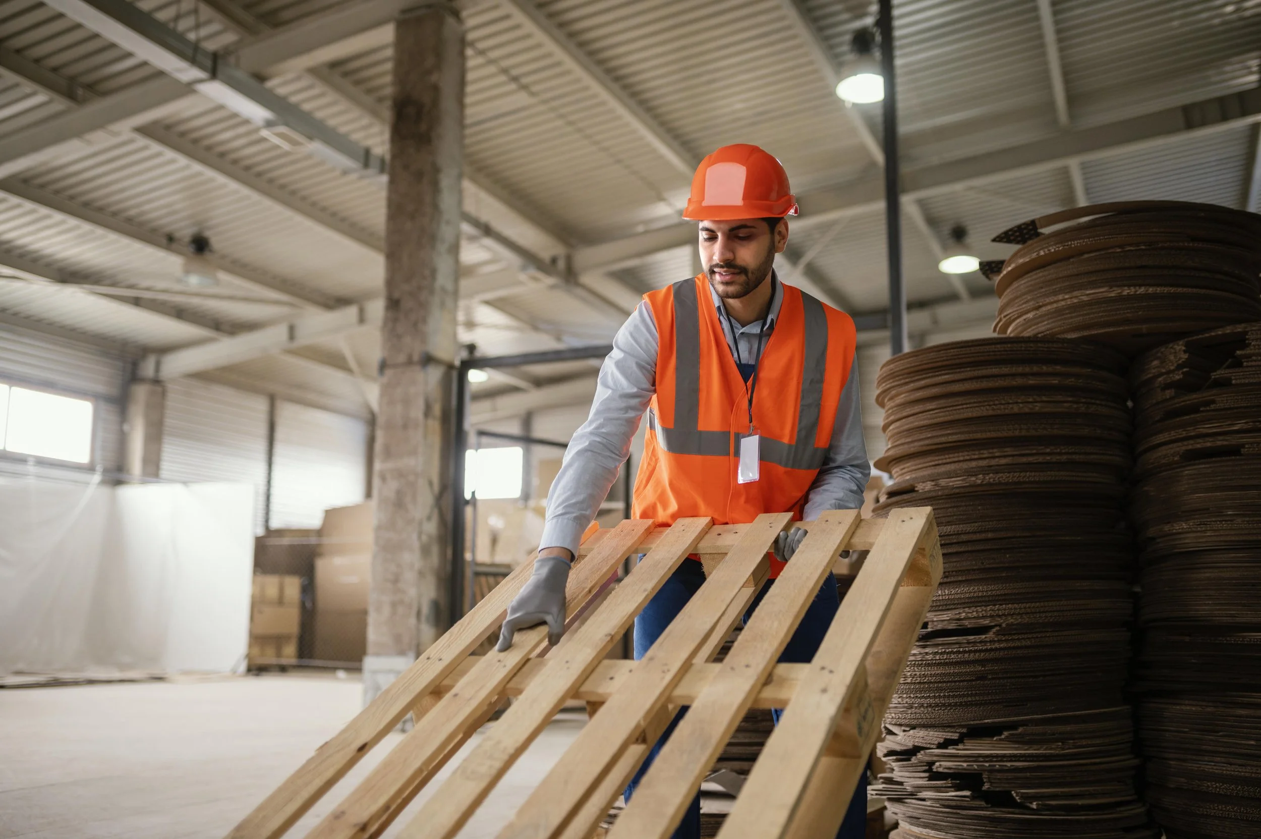 A man wearing a red hard hat and orange safety vest inspecting wooden pallets inside an industrial warehouse with stacks of cardboard sheets nearby.