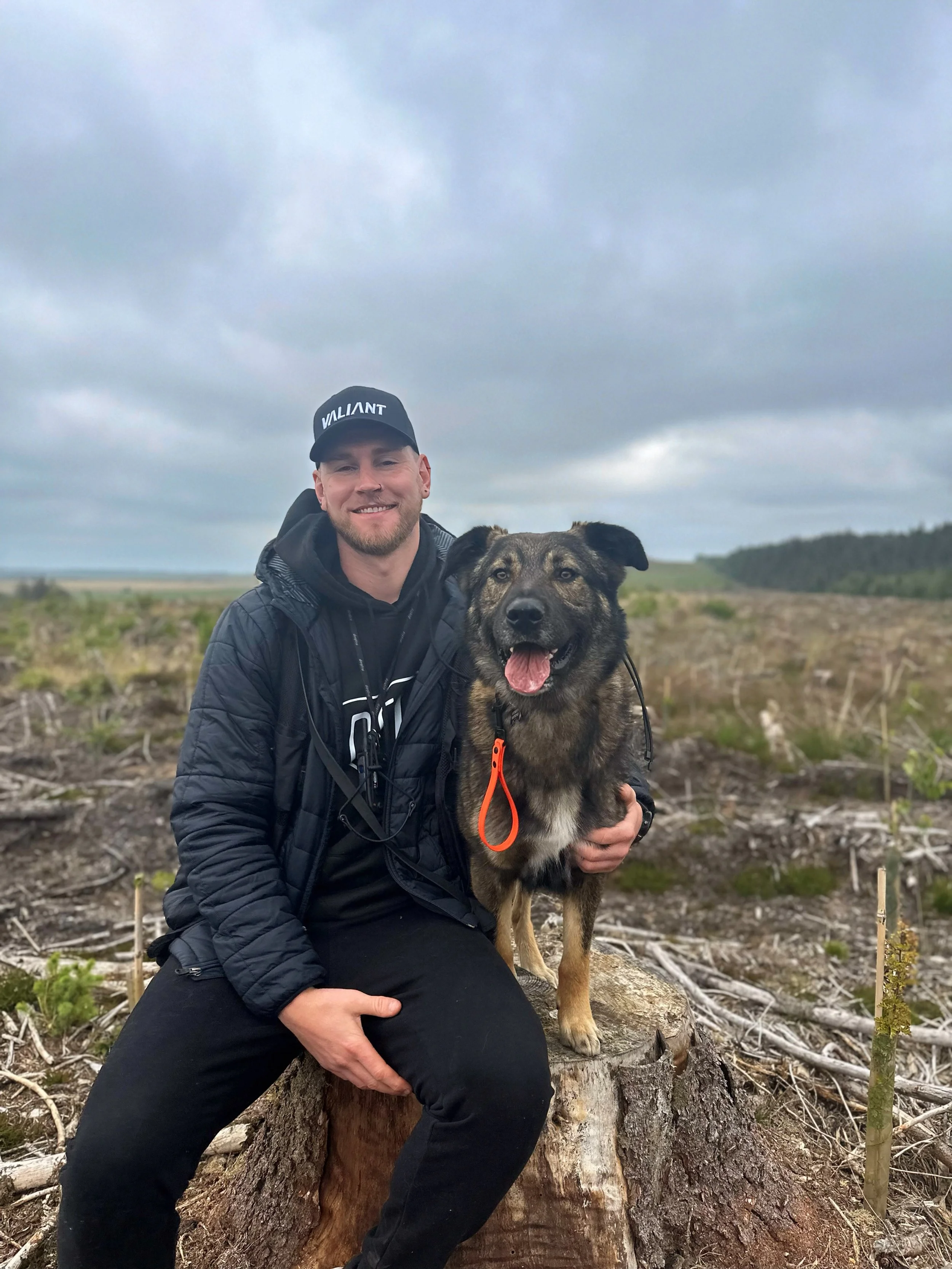 A man in black clothing and a black cap labeled 'VALIANT' sitting on a tree stump beside his dog in an outdoor landscape with cloudy sky, short grass, and trees in the distance.
