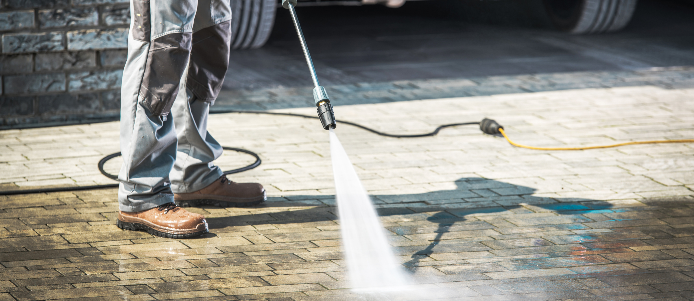 Person spraying water on a brick pavement using a pressure washer, with part of a truck or large vehicle in the background.