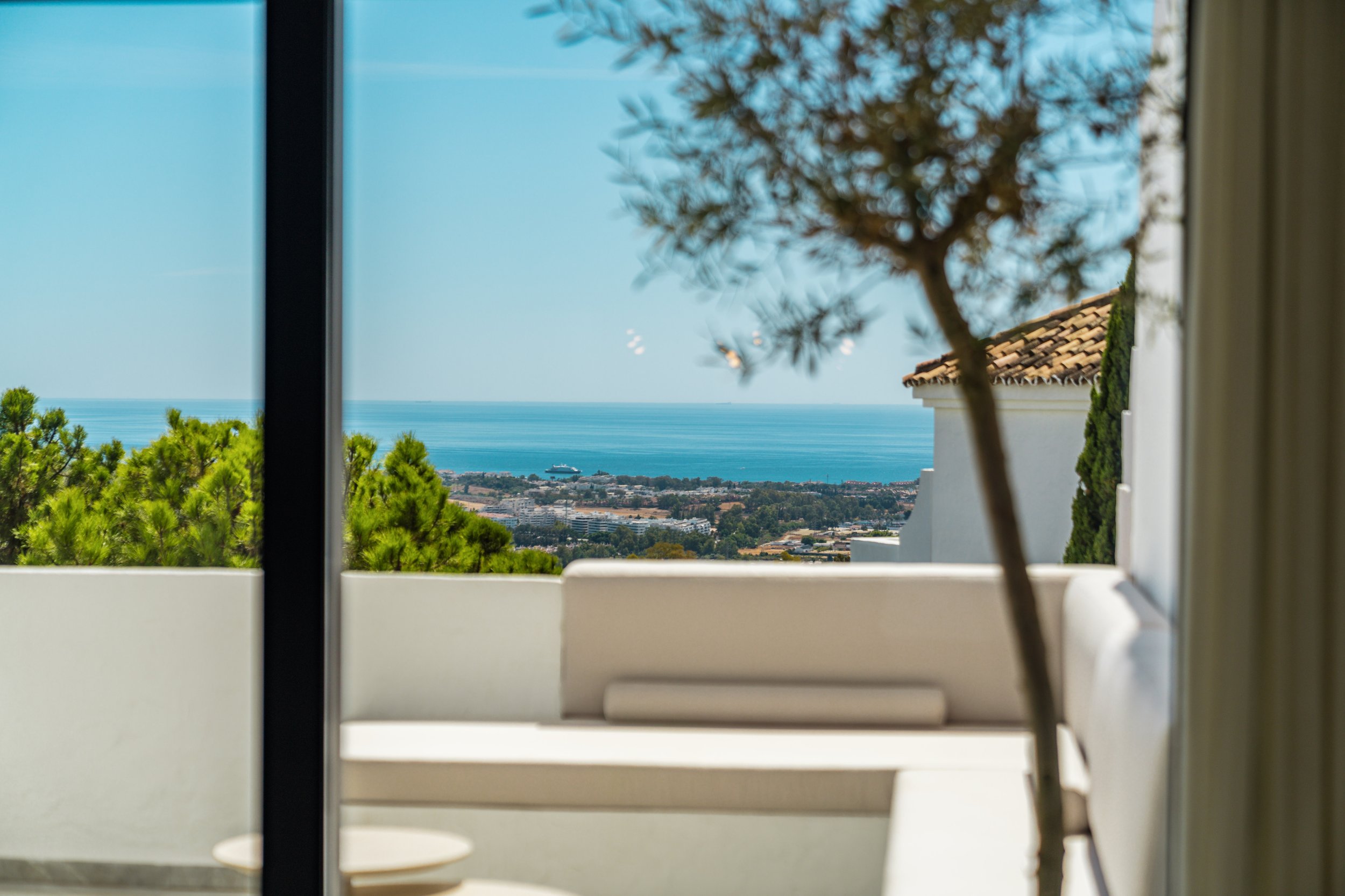 A scene viewed through a window, showing a blue sky, green trees, a tiled roof, and a distant ocean with a ship, partially obscured by window frames and a white balcony.
