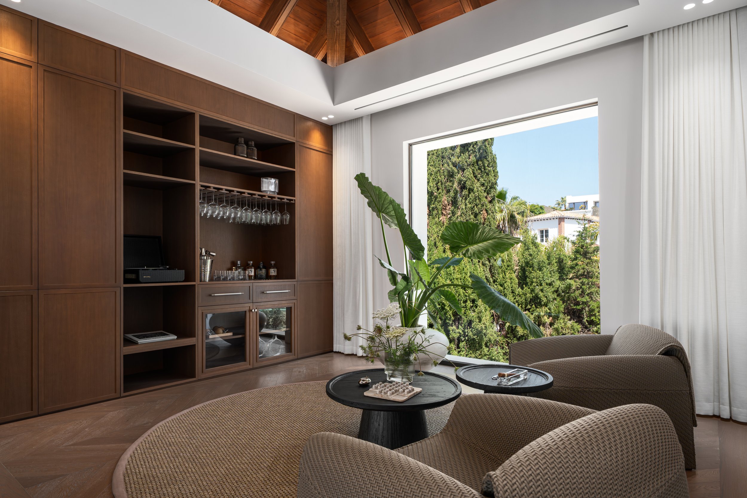 Living room with a large window showing outdoor greenery, wooden wall cabinets, indoor plants, and a sitting area with coffee tables.