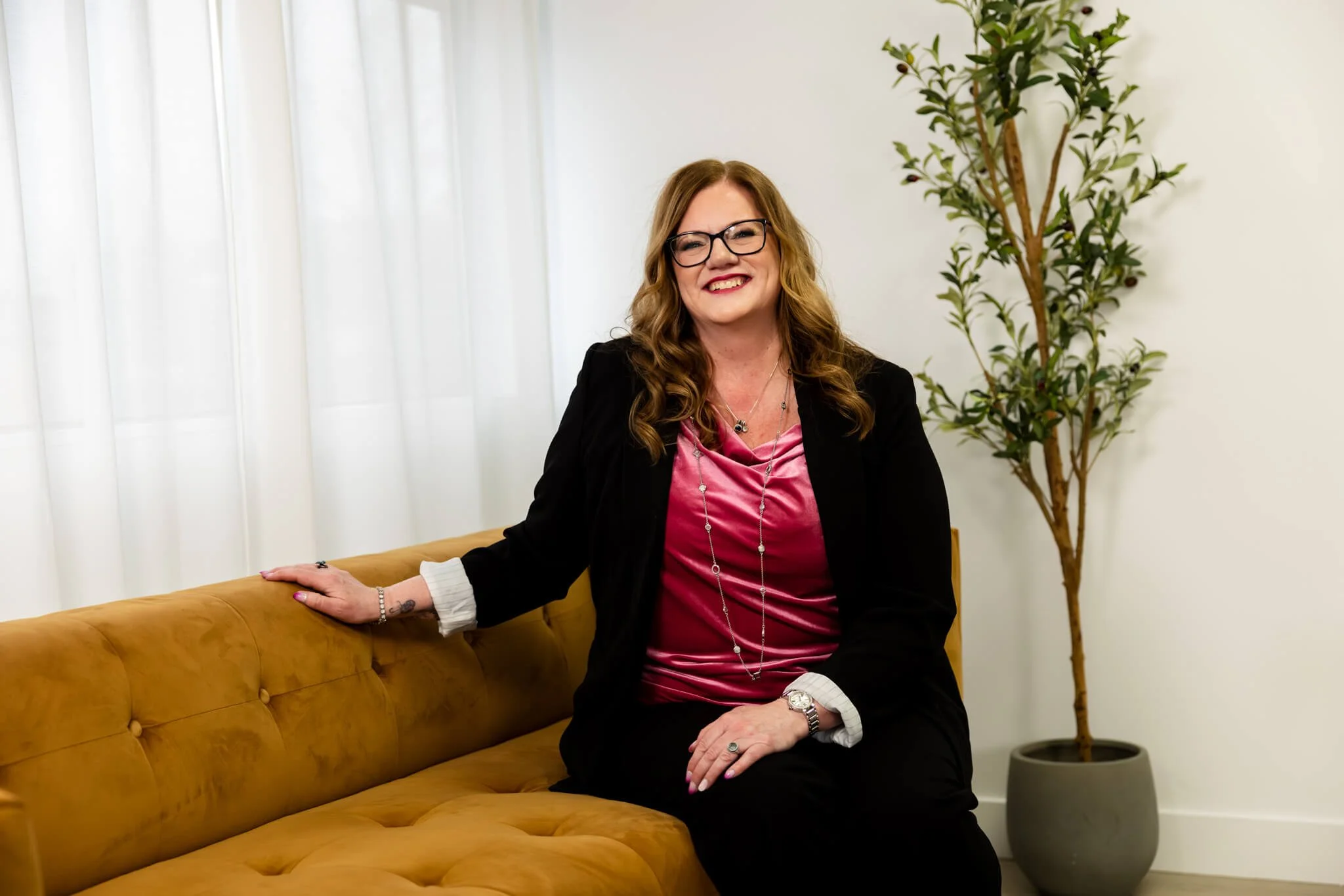 A woman with glasses and long wavy hair smiling, sitting on a mustard-colored sofa in a room with white curtains and a potted tree in the background.