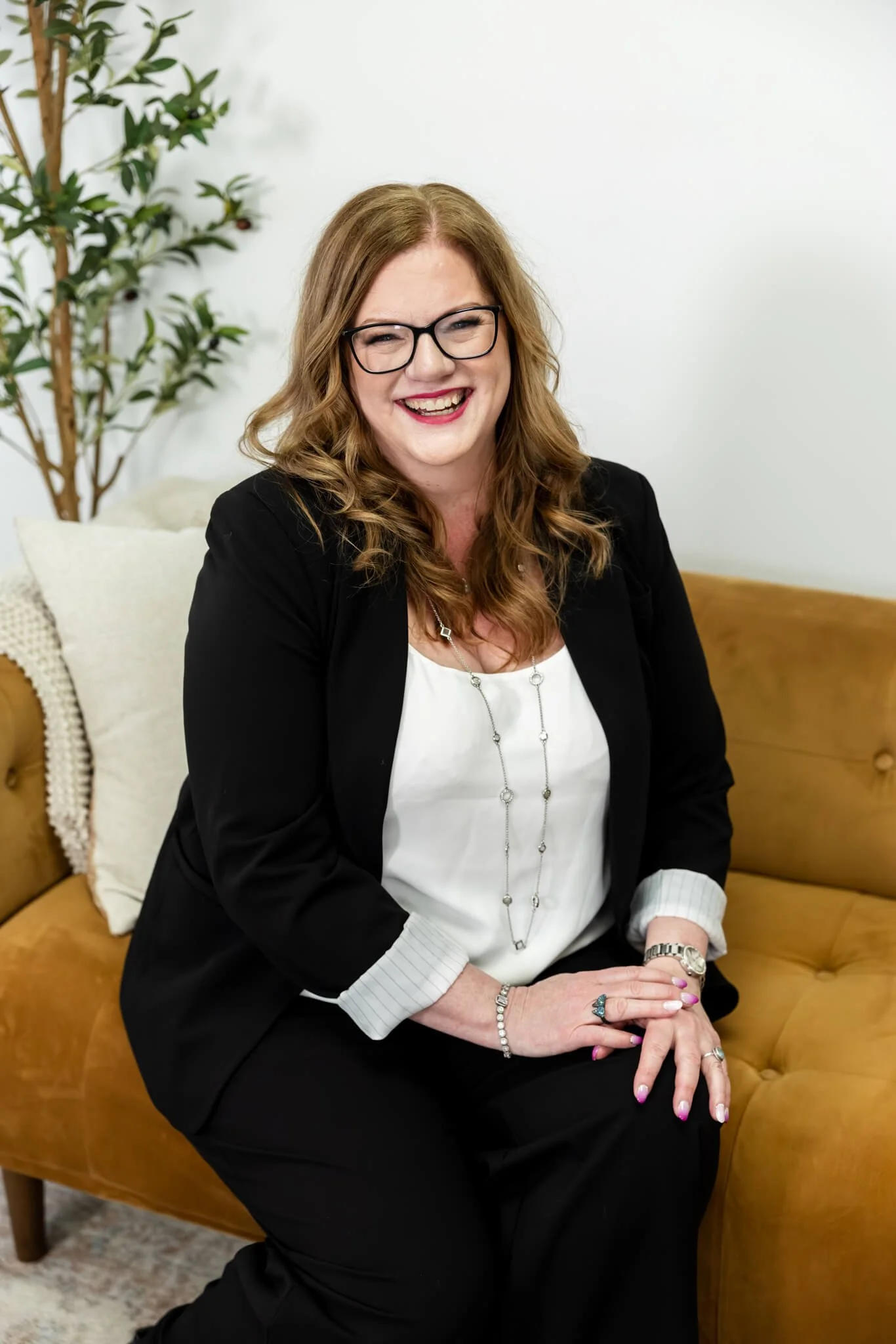 A woman with long curly hair, glasses, and red lipstick sitting on a tan leather couch with a white pillow, dressed in a black blazer and white blouse, smiling at the camera.