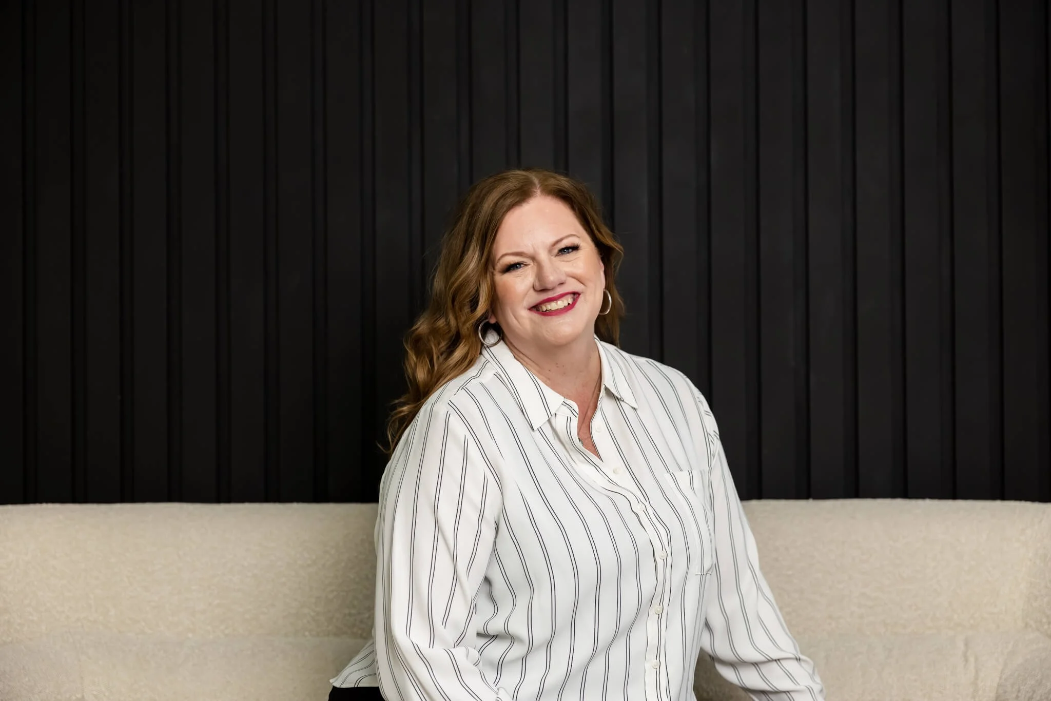 A woman with wavy auburn hair, wearing hoop earrings and a white shirt with black vertical stripes, standing in front of a black paneled wall and beige counter, smiling at the camera.