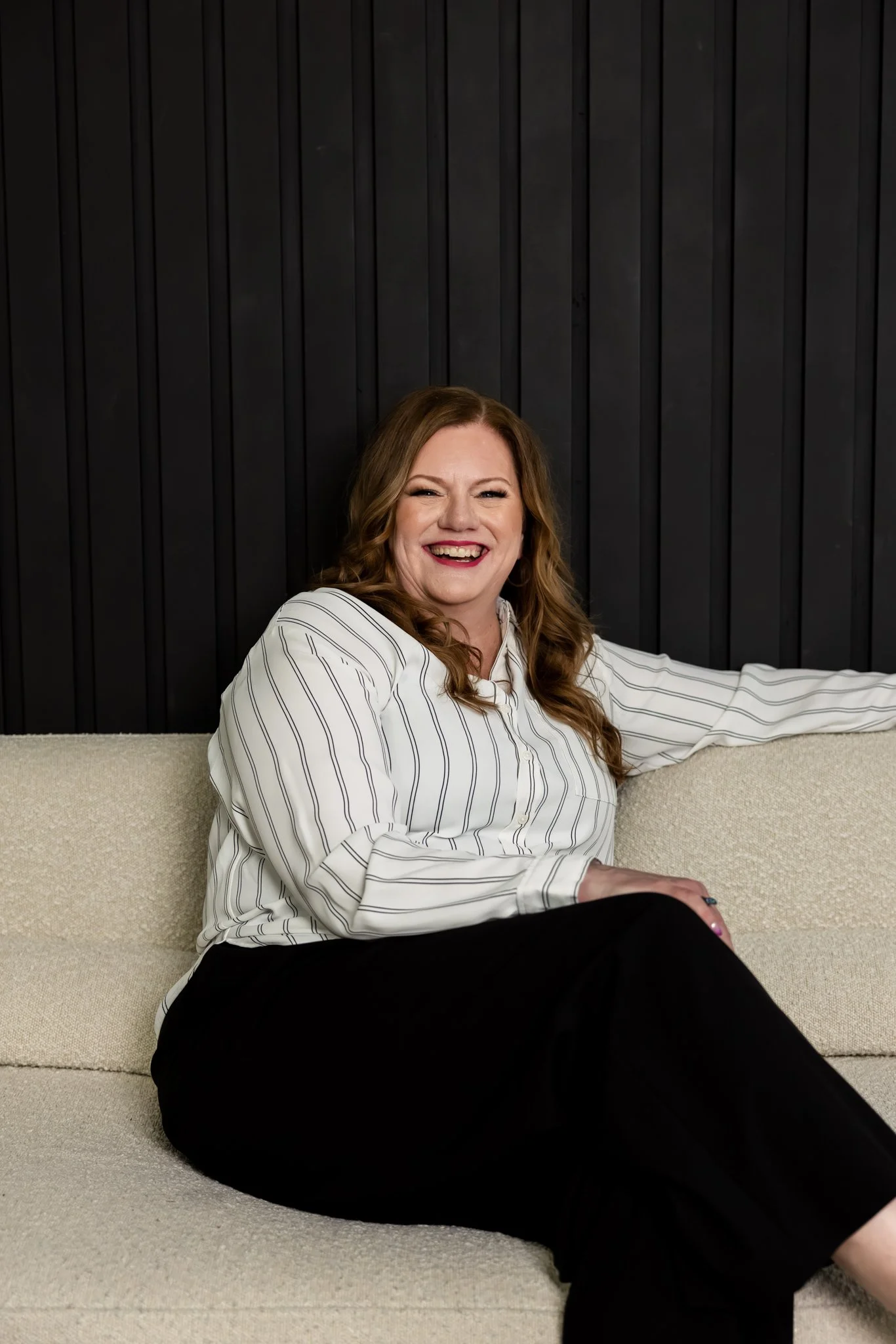 A woman with curly hair is sitting on a light-colored couch, smiling broadly and wearing a white, striped shirt with black pants. She is leaning back with her right arm resting on the back of the couch, in front of a black wooden panel wall.
