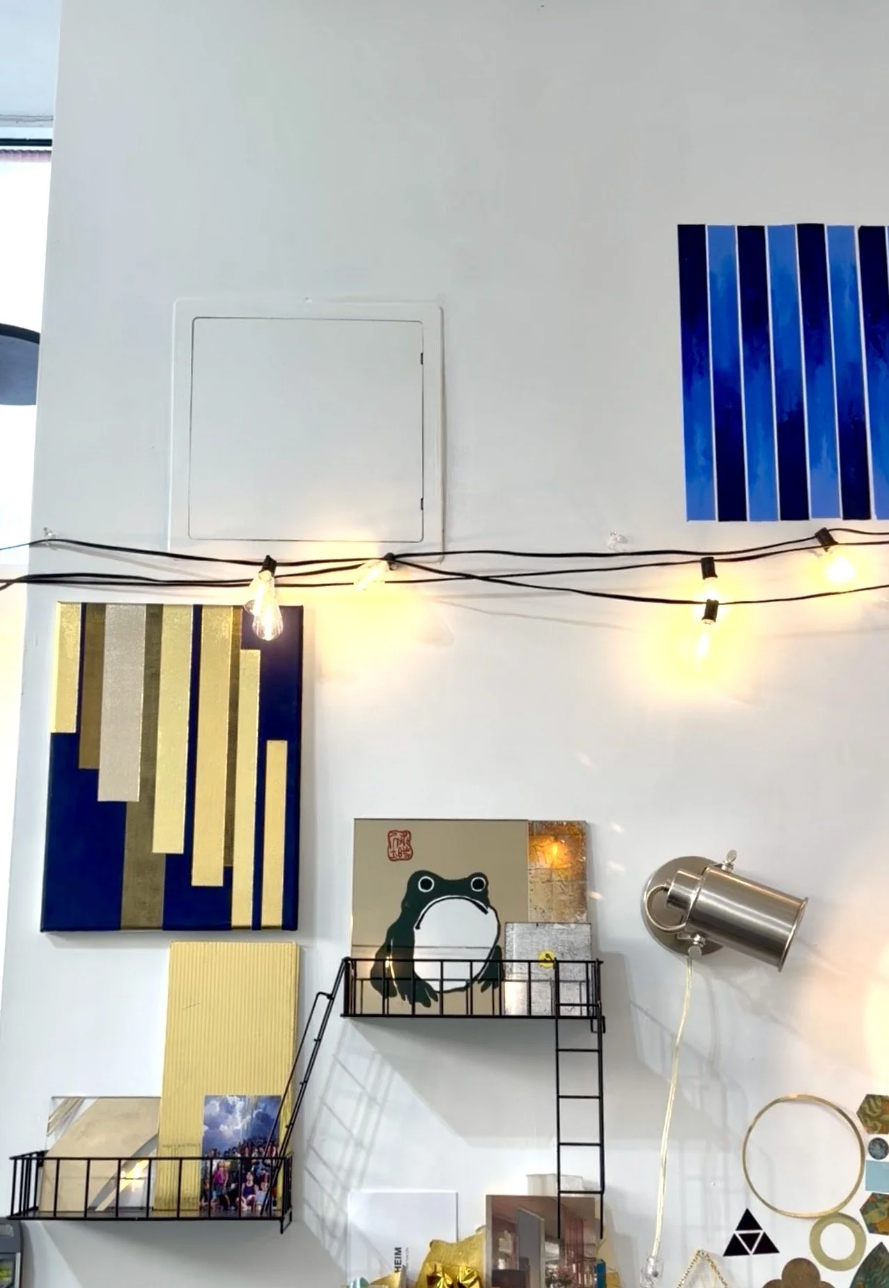 A white wall decorated with abstract art, a metal wall-mounted lamp, string lights, and various small decorative items including a canvas with a frog illustration and geometric decorations.
