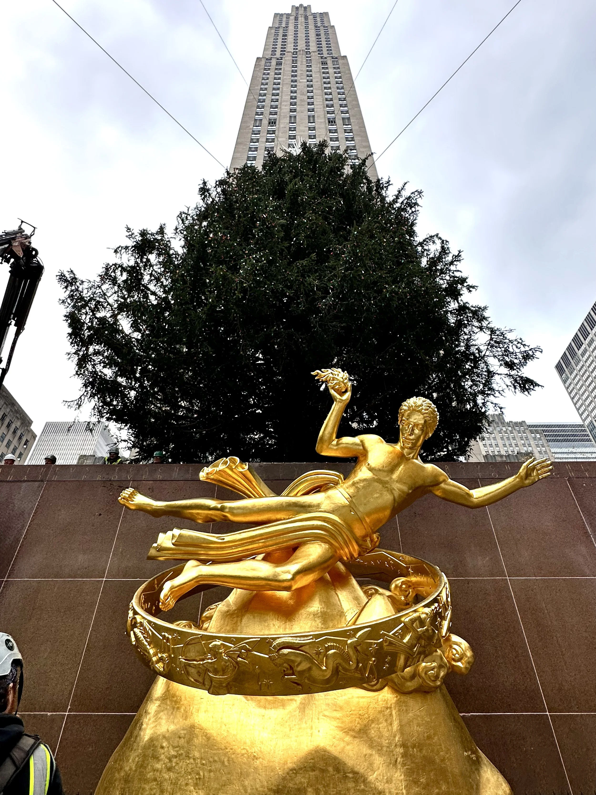 Gold-colored statue of a person lying on a large bell, set against a tall building and a large tree with a cloudy sky.