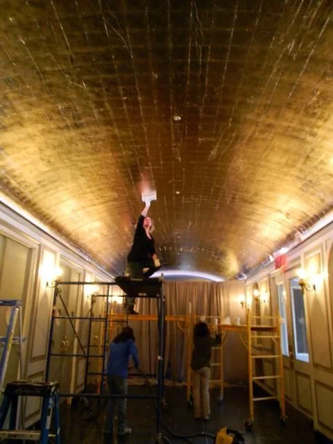 Workers installing or repairing a large gold ceiling in an elegant room with ornate wall paneling and multiple windows.