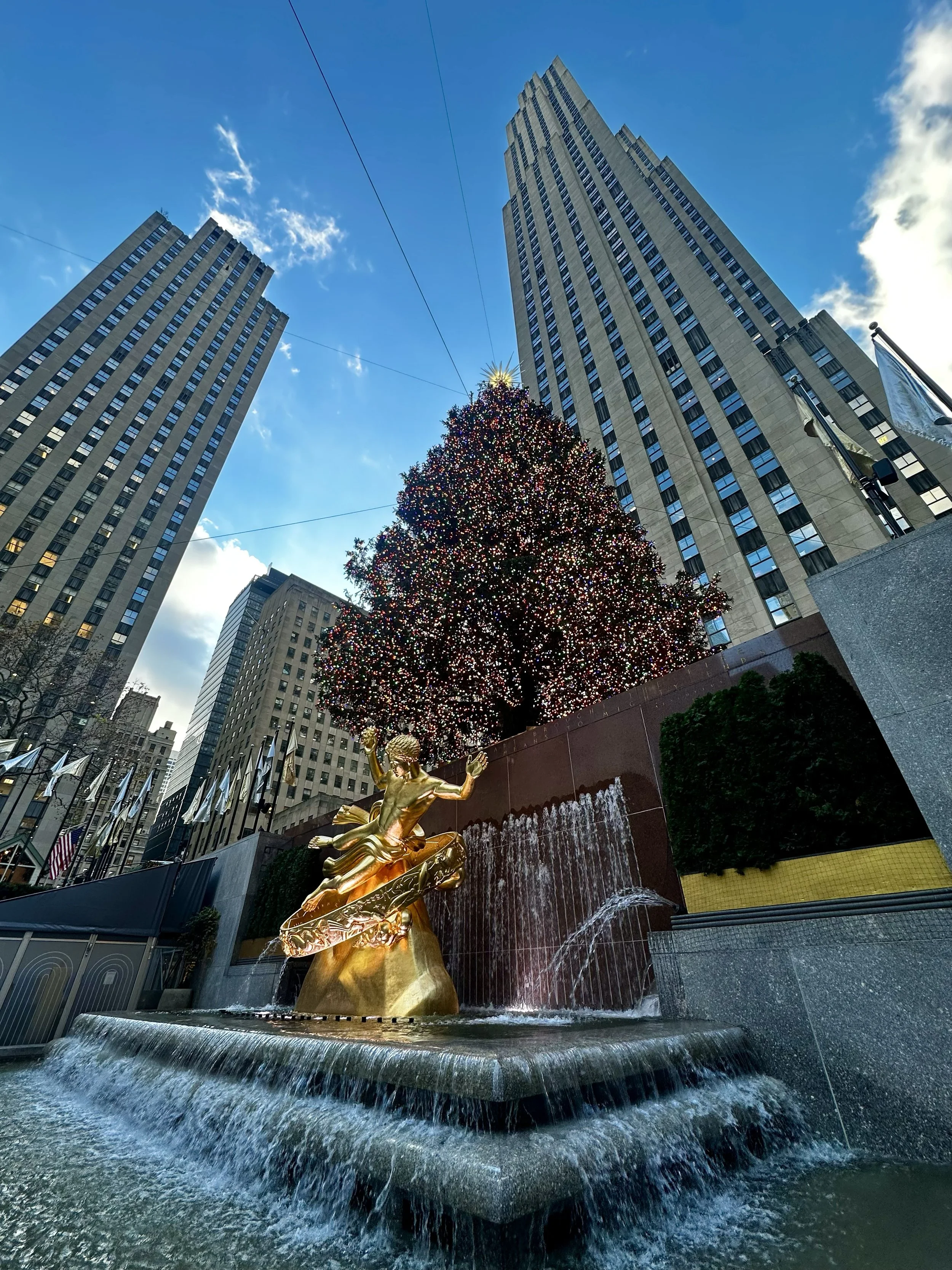 A view of the Rockefeller Center Christmas Tree with a golden statue of Prometheus in the foreground, surrounded by tall buildings in New York City.