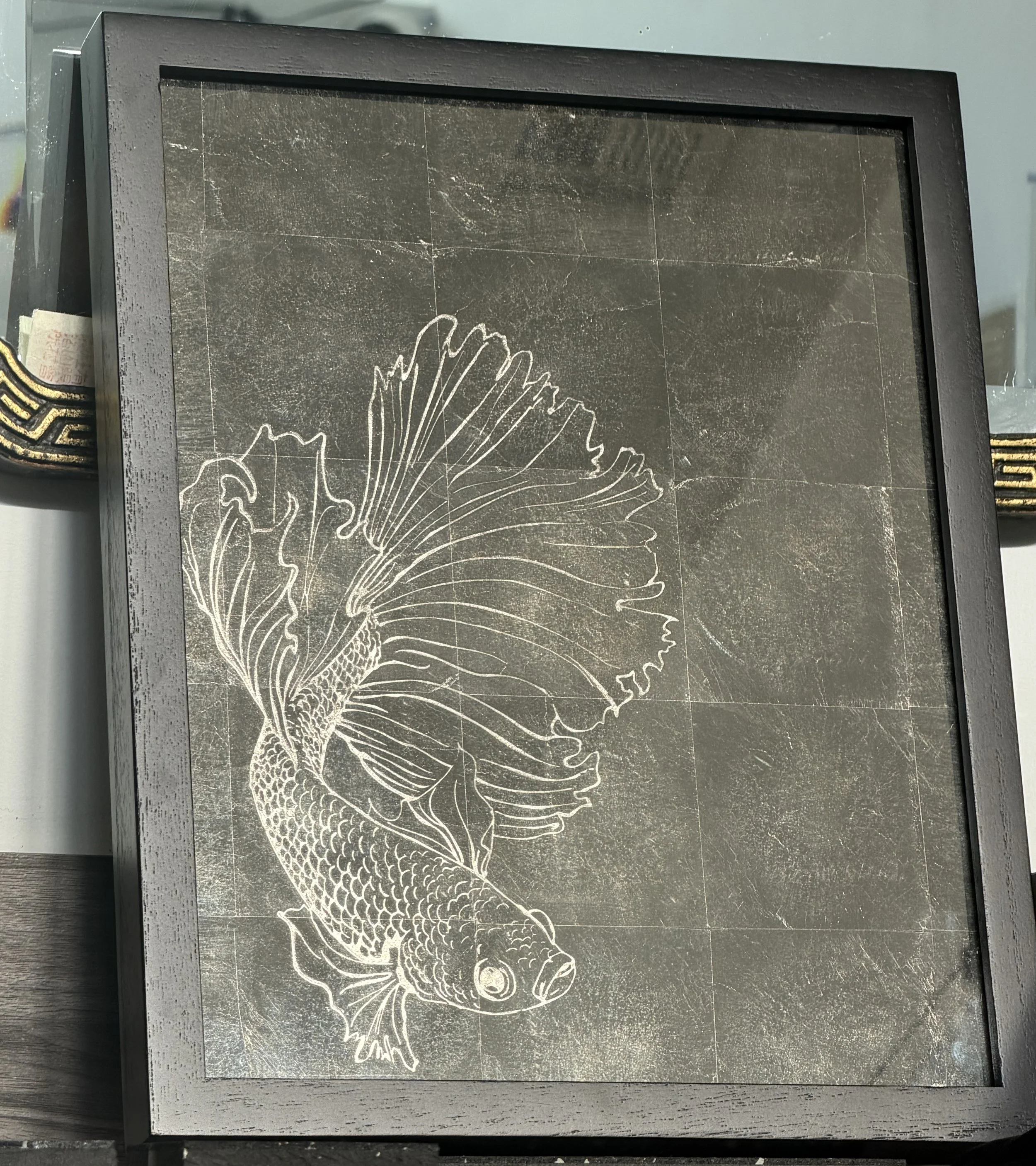 A framed art piece featuring a white line drawing of a fish, likely a betta, on a dark, textured background.