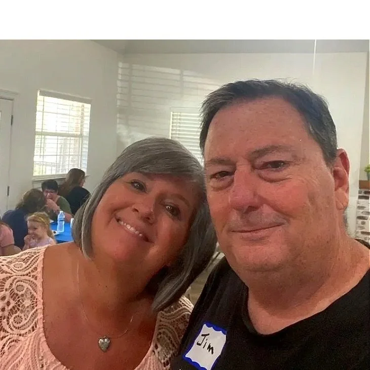 A smiling woman with short gray hair and a necklace, and a man with dark hair and a name tag that says 'Jim', taking a selfie indoors with other people in the background.