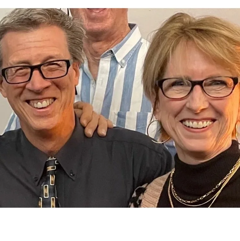 Smiling man and woman wearing glasses, with the woman wearing a black turtleneck and gold jewelry, standing close together, with part of a man's face and shoulder visible behind them, in an indoor setting.