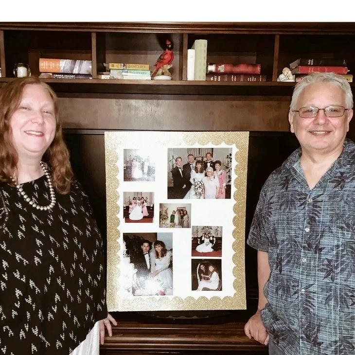 A woman and a man standing on either side of a collage of photographs, with a wooden bookcase in the background.