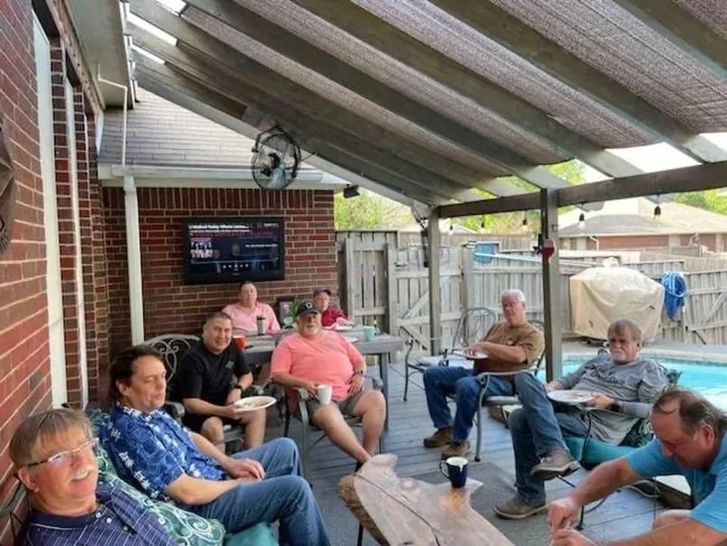 Group of eight adults sitting and standing on a backyard patio, some eating and drinking, under a slanted roof with string lights, a large grill covered with a cover, a wooden fence, a pool, and a television mounted on a brick wall.
