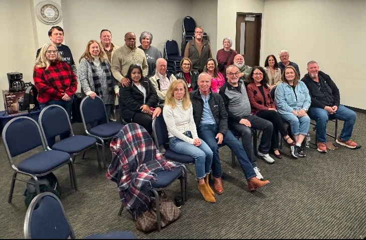 Group of 19 people, five seated in the front row and 14 standing behind them, inside a room with beige walls, dark carpet, and a clock on the wall.