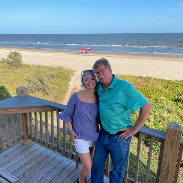 A couple standing on a wooden balcony overlooking a beach with sand and the ocean, a dog, and red umbrellas in the distance.