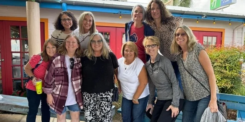 A group of ten women standing outdoors in front of a colorful building with pink doors and green bushes, smiling for the photo.