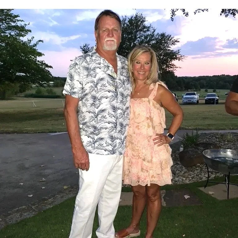 A man and woman standing outdoors during sunset, smiling at the camera. The man is wearing a short-sleeved, button-up shirt with a leaf pattern and white pants. The woman is wearing a light pink, ruffled summer dress with sandals.