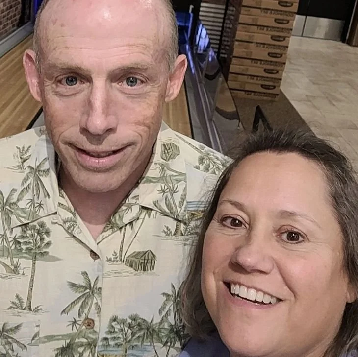 A smiling middle-aged woman and a middle-aged man with short light hair taking a selfie together in what appears to be a store or shopping area.