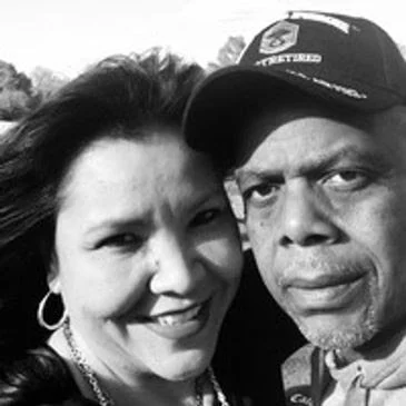 A black-and-white photo of a smiling woman and a man wearing a baseball cap, outdoors.