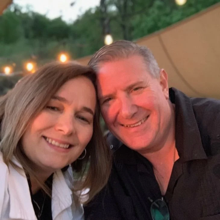 A smiling woman with shoulder-length light brown hair and a man with short gray hair are close together, taking a selfie outdoors during evening time, with blurred string lights in the background.