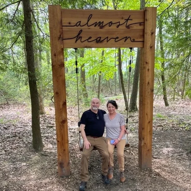 A man and woman sitting on a wooden swing in a forest, with a wooden sign above them that says 'almost heaven'.