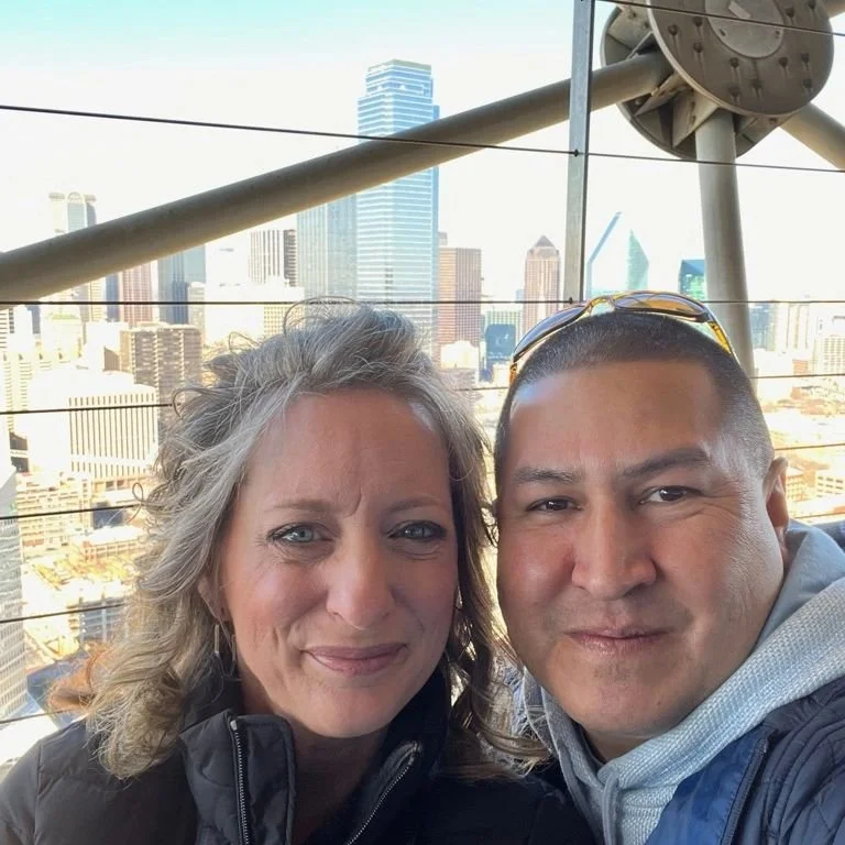 A woman and a man taking a selfie on a rooftop with a city skyline in the background, featuring skyscrapers and tall buildings.