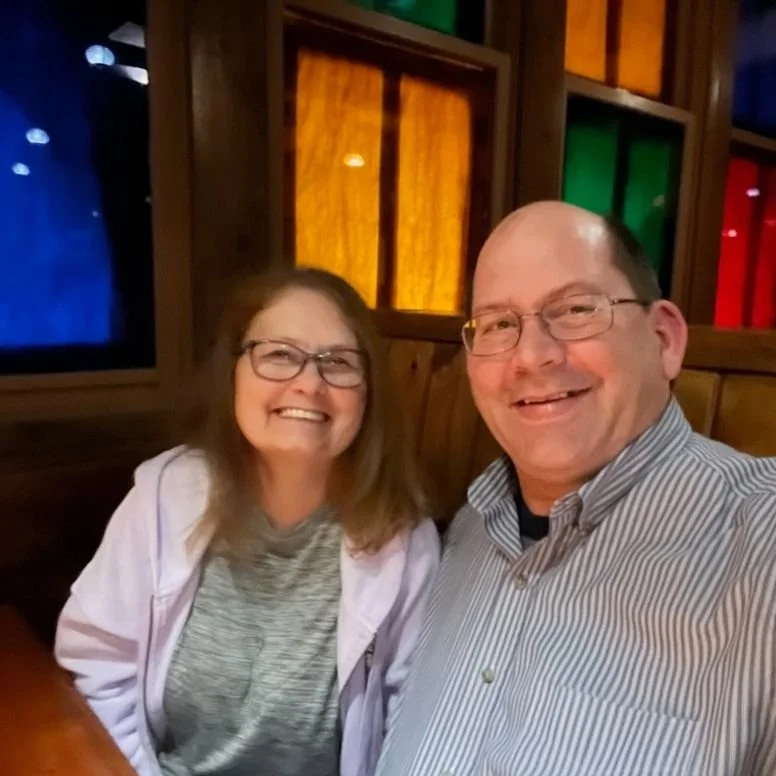 A smiling man and woman taking a selfie inside a restaurant with colorful stained glass windows in the background.