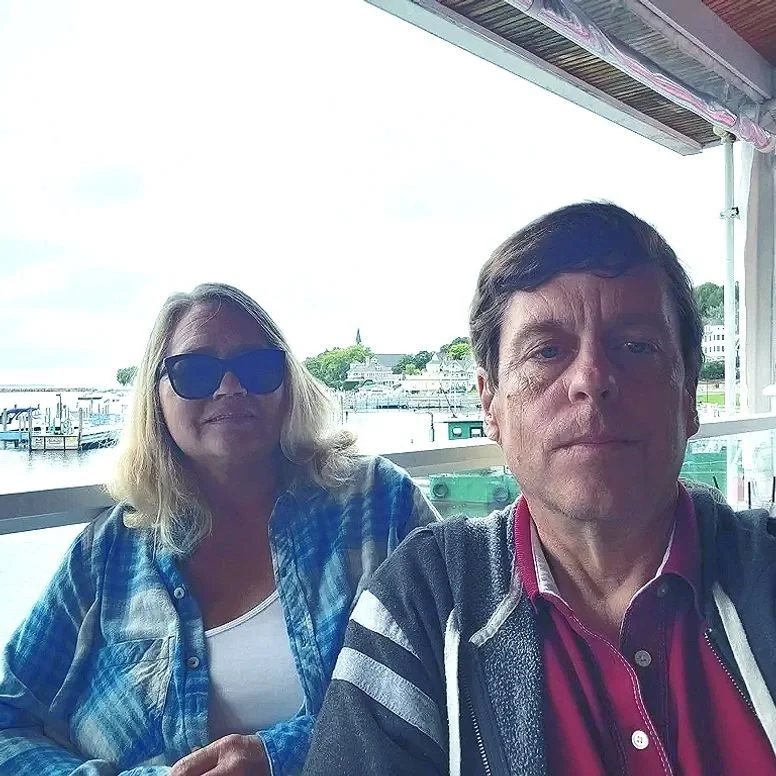 A man and a woman taking a selfie on a boat, with a view of a marina and buildings in the background. The woman is wearing sunglasses and a blue plaid shirt, while the man is wearing a red shirt and a dark jacket.
