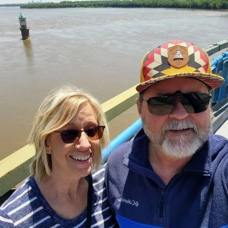 A smiling older woman and man taking a selfie on a bridge over a river, with a small lighthouse in the water behind them. The man is wearing a colorful cap and sunglasses; the woman is wearing sunglasses and a striped shirt.