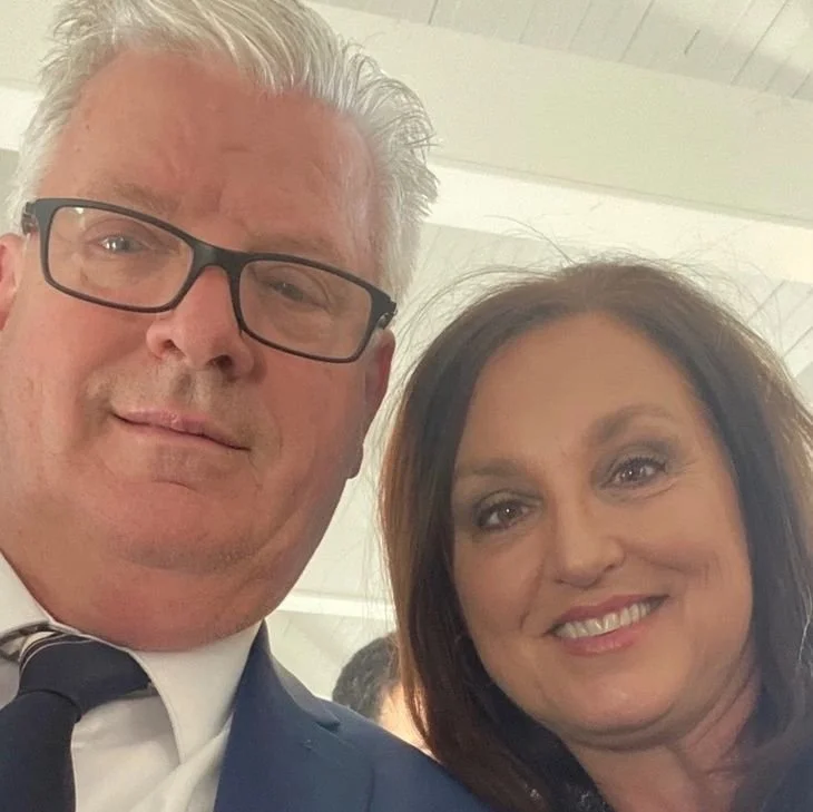 A man with gray hair, black glasses, and light skin, wearing a suit and tie, posing for a selfie with a woman with shoulder-length brown hair, makeup, and light skin, smiling at the camera.