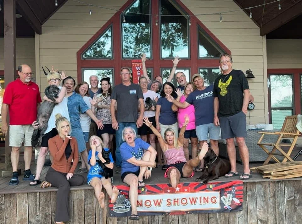 Group of people on a wooden deck in front of a house with large triangular windows, posing for a picture with some raising their hands and smiling, some sitting on the deck, and a dog among them. There is a sign that reads "NOW SHOWING" in front of t