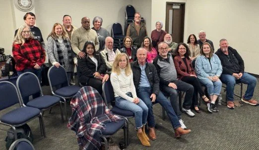 Group of people sitting and standing in a room with empty chairs, posing for a photo.