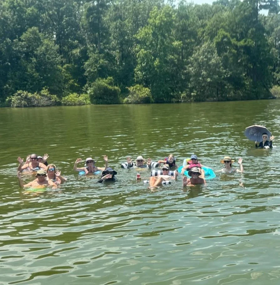Group of people floating and waving in a lake with trees in the background, some wearing hats and sunglasses, and one person with an umbrella.