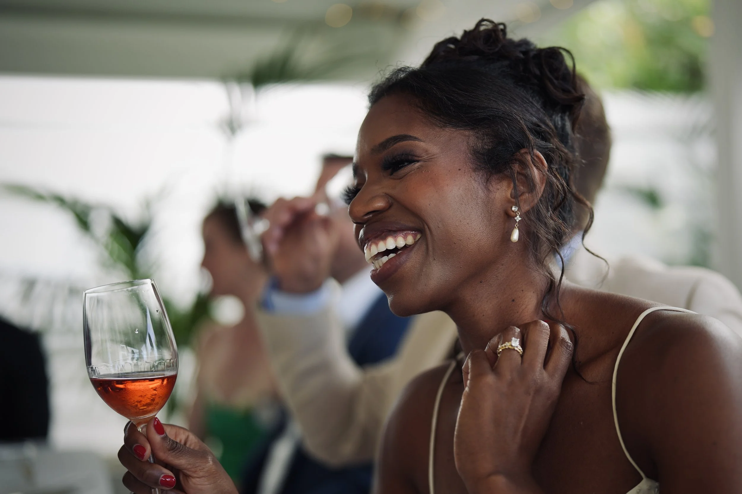 A woman smiling while holding a wine glass with rosé wine at a social gathering, with other people in the background.
