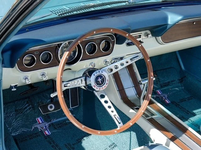 Interior of a vintage Ford Mustang car, showing the steering wheel, dashboard with gauges, and part of the floor with floor mats.
