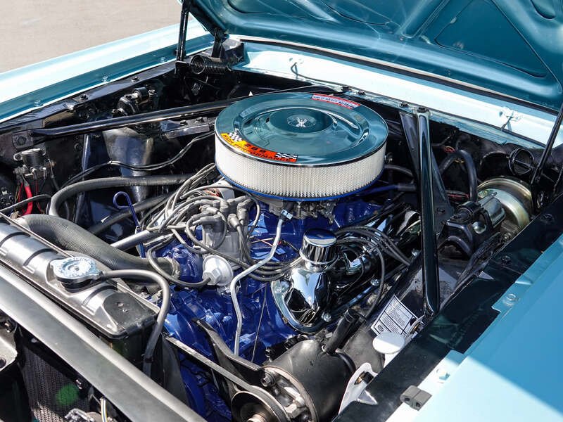 Inside view of a car engine bay showing a V8 engine with a round air cleaner on top, various hoses, wires, and metallic components.