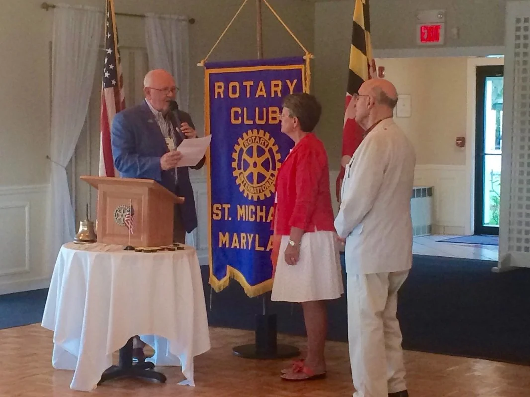 A man in a blue blazer reads from a paper at a podium while two people, a woman in a red blazer and a man in a white suit, stand facing him during a Rotary Club event in Maryland. Behind them are American and Maryland flags and a Rotary Club banner.