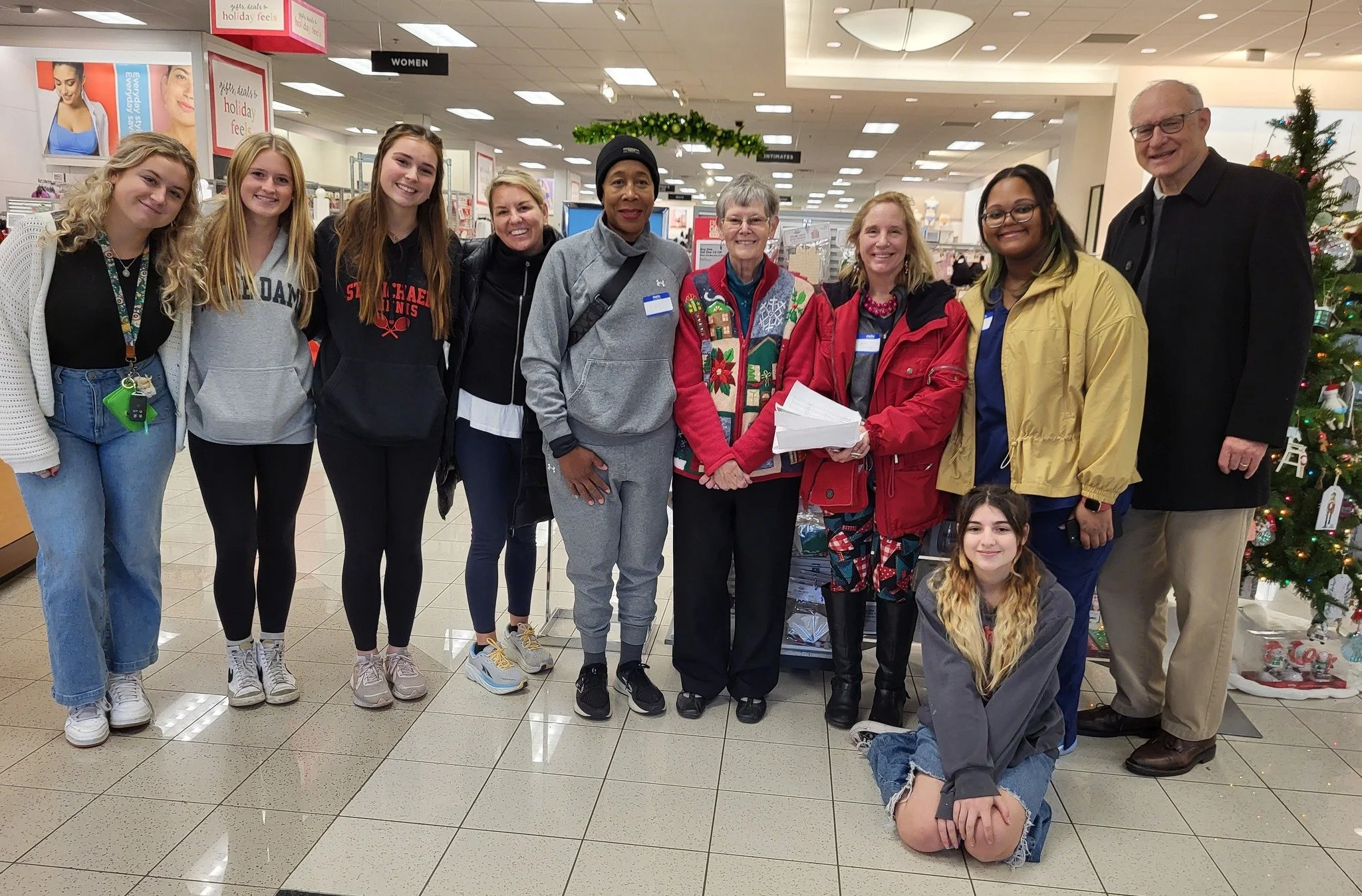 Group of ten diverse people standing together inside a store near a decorated Christmas tree, with some holding papers and others smiling at the camera.
